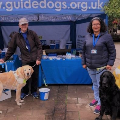 Two volunteers from the Exeter group stand by a stall with Guide Dogs merchandise and two dogs.