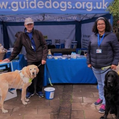 Two volunteers stand by a fundraising stall with Guide Dogs merchandise in the background. They are 